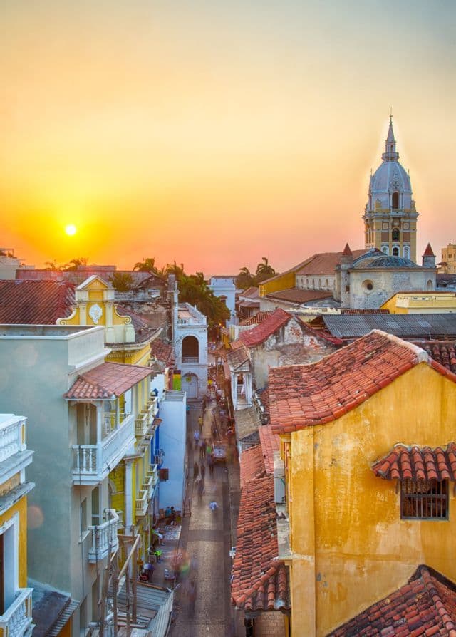Eine erhöhte Ansicht einer Stadtstraße mit farbenfrohen Gebäuden und roten Ziegeldächern bei Sonnenuntergang, mit einer Kathedrale im Hintergrund.