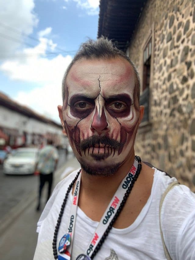 Un uomo con il viso dipinto da teschio dettagliato e un laccetto WeRoad posa per un selfie su una strada di ciottoli.