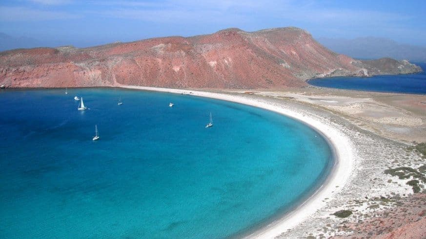 Una veduta aerea di barche a vela su acqua turchese in un'ampia baia accanto a una spiaggia di sabbia bianca a mezzaluna e colline rosse.