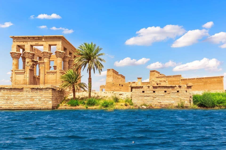 An ancient stone temple with large columns and palm trees on the bank of a river under a blue sky with clouds.