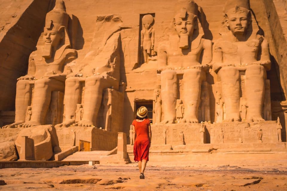 Eine Frau in einem roten Kleid und Strohhut geht auf einen großen, antiken Tempel zu, der in eine Klippe mit kolossalen sitzenden Statuen gemeißelt ist.