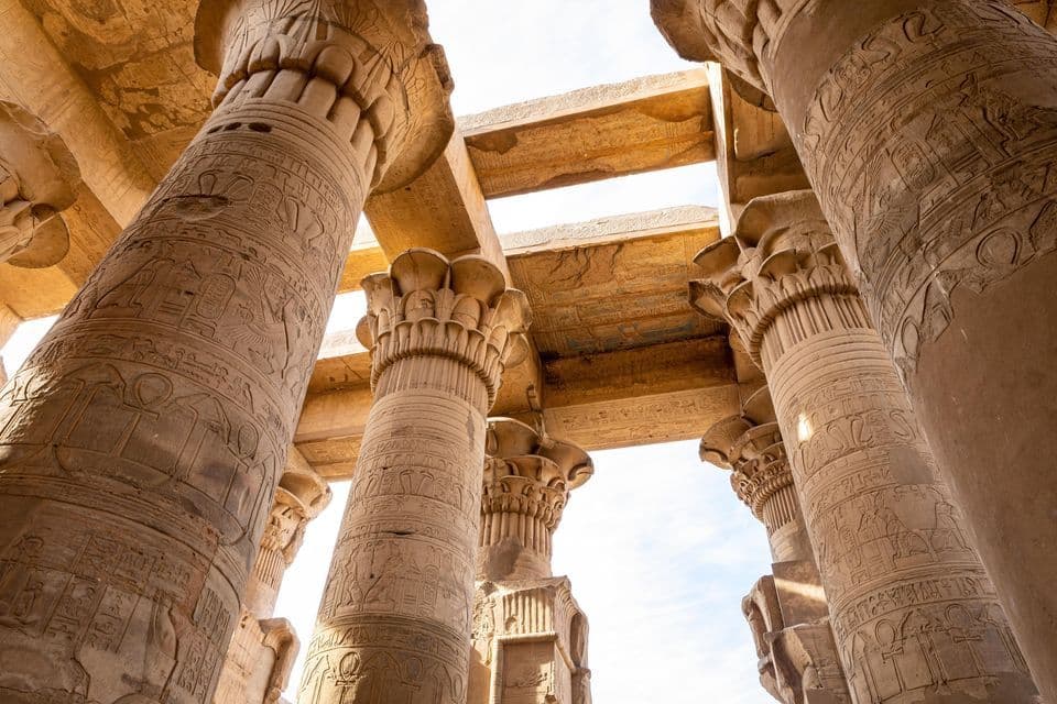 A low-angle view of large stone columns with carved hieroglyphics inside an ancient temple, supporting a stone ceiling.