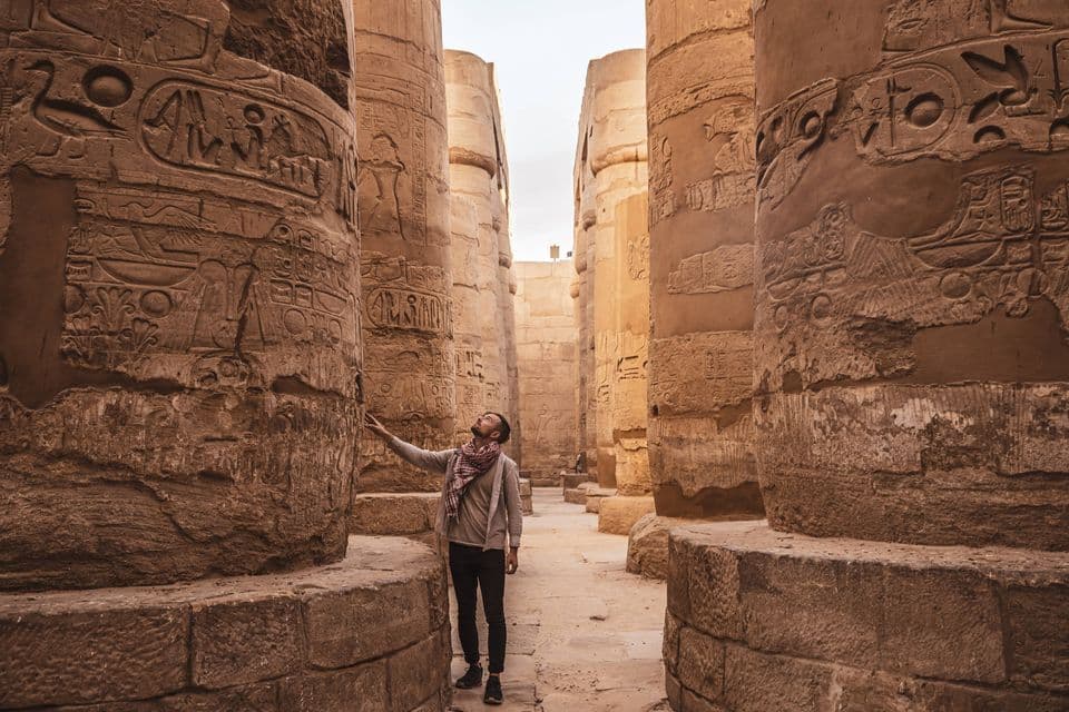 Ein Mann steht in einer alten Tempelhalle und blickt zu den Hieroglyphen auf, die in die massiven Steinsäulen gemeißelt sind.