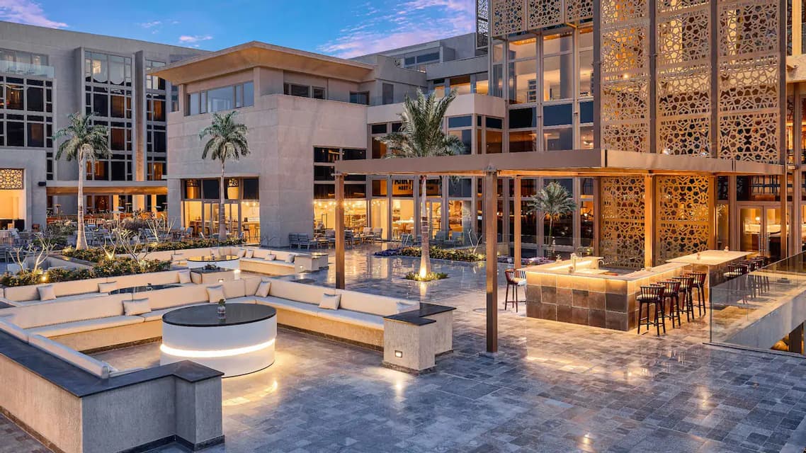 Una terrazza esterna di un hotel moderno al tramonto, con aree salotto illuminate, un bar e palme.