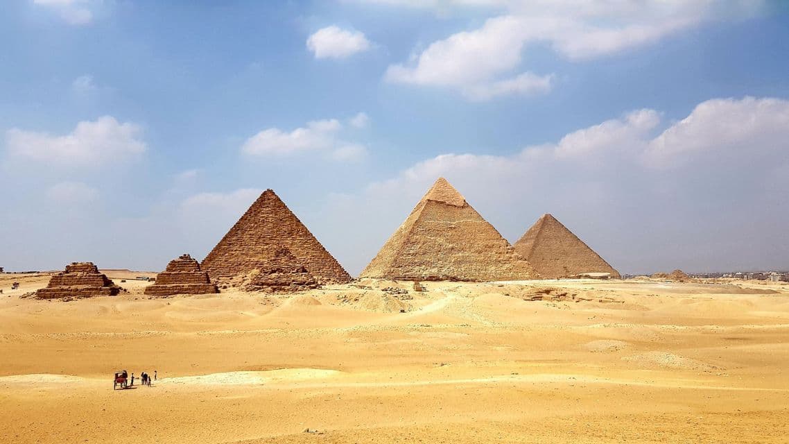 A wide view of several large and small pyramids in a vast desert under a blue sky with scattered clouds, with a carriage in the distance.
