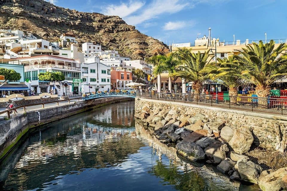 A sunlit canal runs through a coastal town with white buildings and palm trees, set against a rocky hillside under a blue sky.