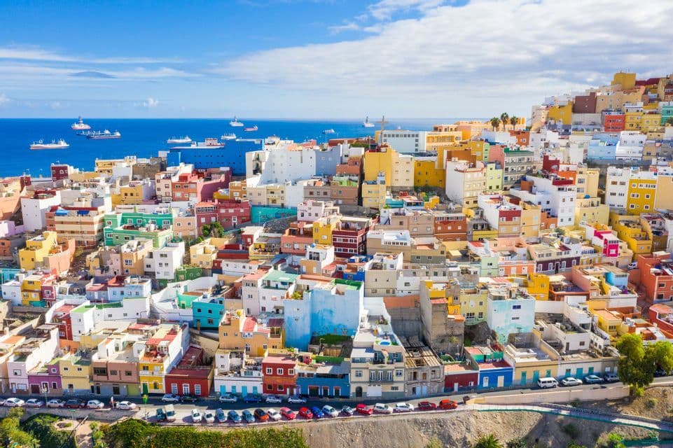 An aerial view of a colorful hillside city overlooking a blue ocean with ships under a partly cloudy sky.