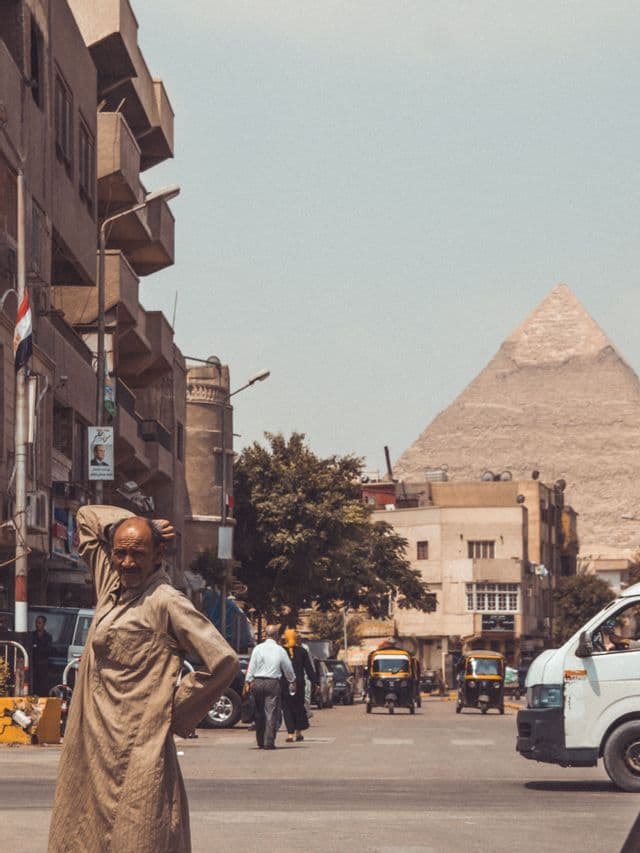 Un homme en tunique marron clair se tient debout dans une rue animée, avec du trafic, des bâtiments et une grande pyramide en arrière-plan.