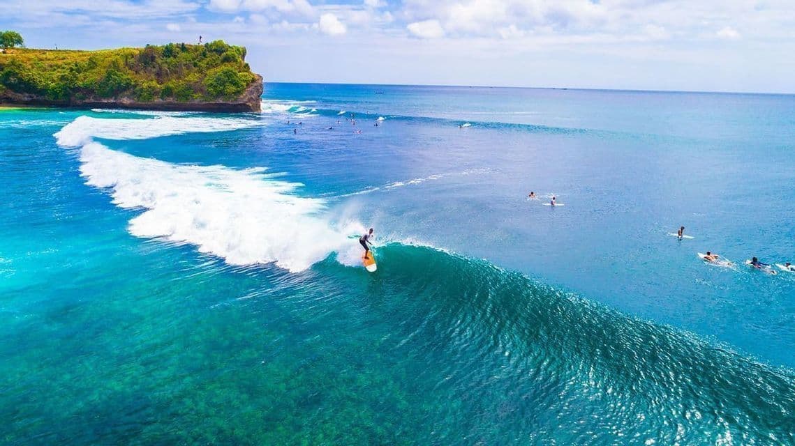 An aerial view of a surfer riding a large wave in turquoise ocean water near a lush green cliff.