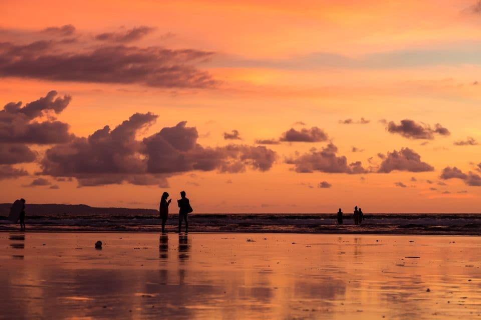 Silhouettes d'un voyage en groupe WeRoad sur une plage mouillée au coucher du soleil, avec un ciel orange et des nuages se reflétant dans le sable.