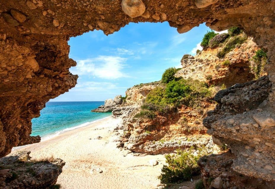 A view from inside a rock archway overlooking a sandy beach, turquoise sea, and cliffs under a blue sky.