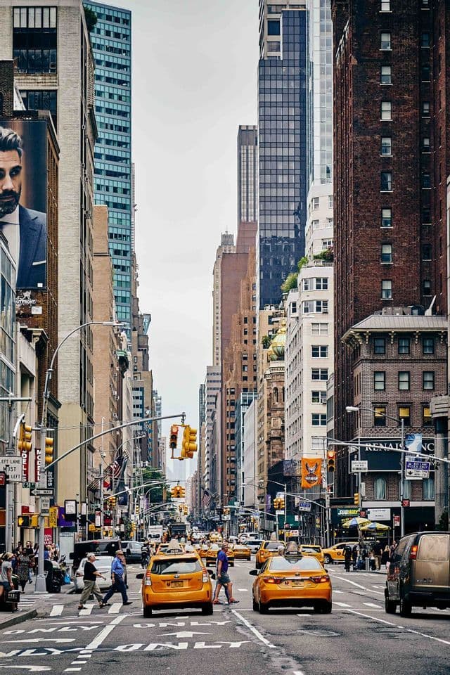 A busy city street with yellow taxis driving and pedestrians crossing, flanked by tall skyscrapers under a clear sky.