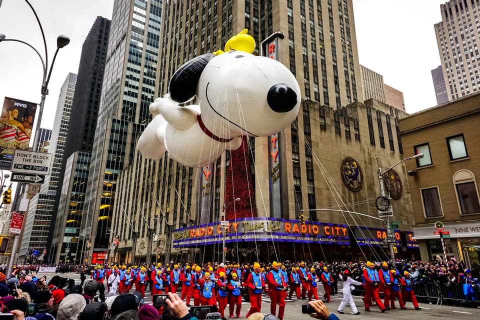 Un grande pallone di Snoopy fluttua sopra una parata in una strada cittadina di fronte alla Radio City Music Hall, con la folla che osserva.
