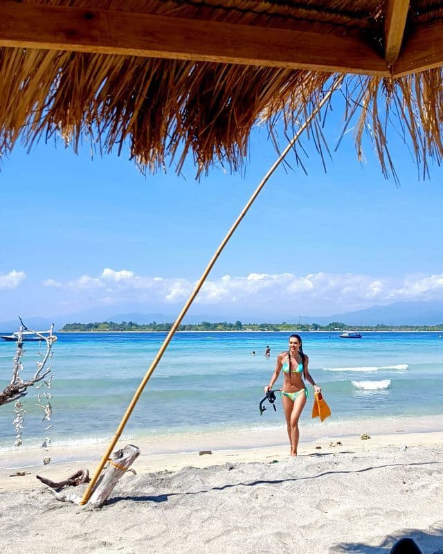 Une femme en bikini tenant un équipement de snorkeling marche sur une plage de sable aux eaux turquoises, vue de sous un toit de chaume.