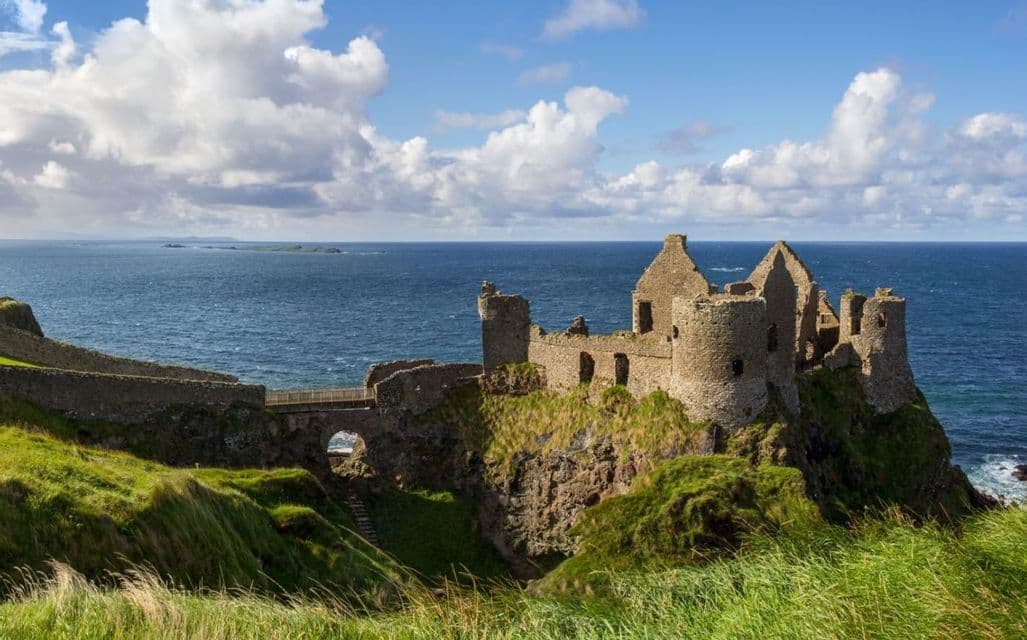 Le rovine di un castello in pietra si stagliano su una verde e rocciosa scogliera a picco sull'oceano, sotto un cielo parzialmente nuvoloso.