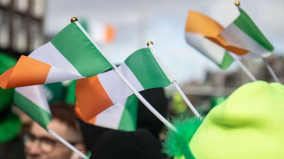 Primo piano di diverse piccole bandiere irlandesi su aste bianche sollevate in mezzo a una folla, con una persona con un cappello verde visibile.