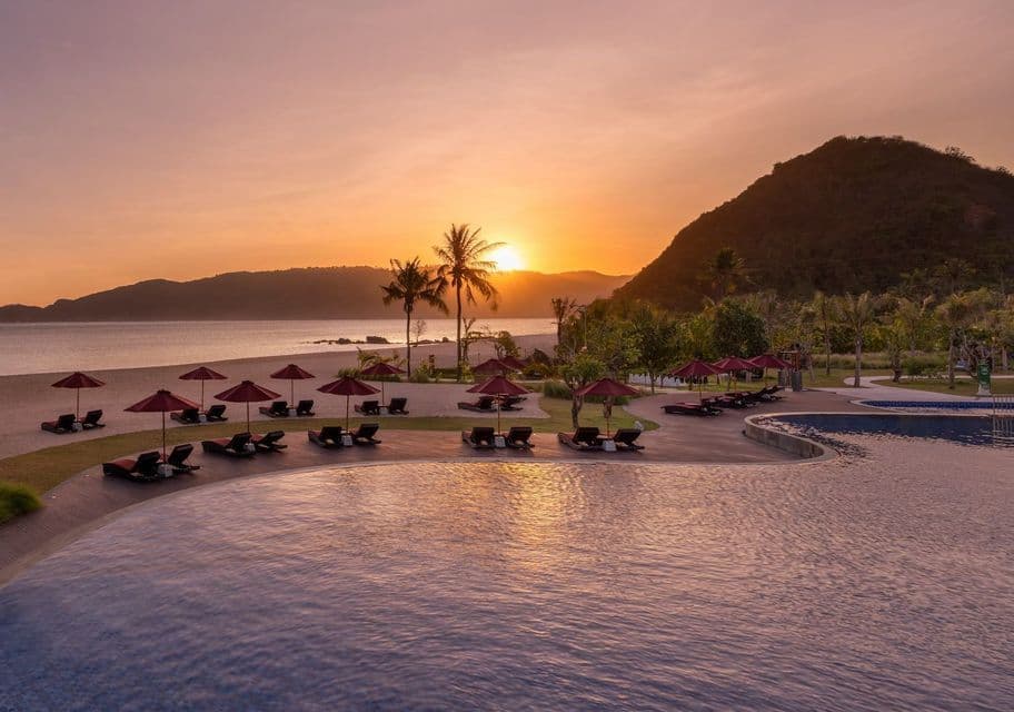La piscina di un resort si affaccia su una spiaggia sabbiosa con lettini e ombrelloni rossi, mentre il sole tramonta sull'oceano e sulle colline.