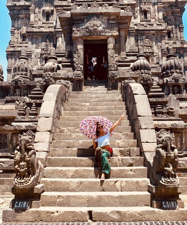 Una donna con un ombrello variopinto è seduta sugli ampi gradini in pietra di un antico tempio riccamente decorato.
