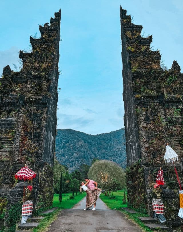 Una coppia posa su un sentiero tra due imponenti pilastri di pietra ricoperti di muschio, con montagne boscose sullo sfondo.