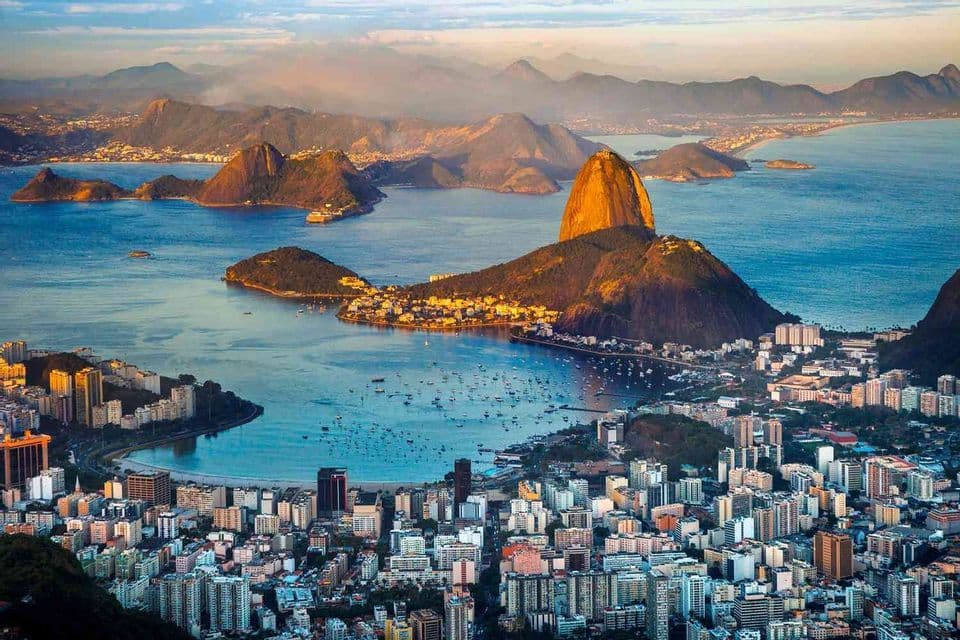 An aerial view of a sprawling coastal city and bay with boats, nestled among prominent mountains rising from the sea during sunrise.