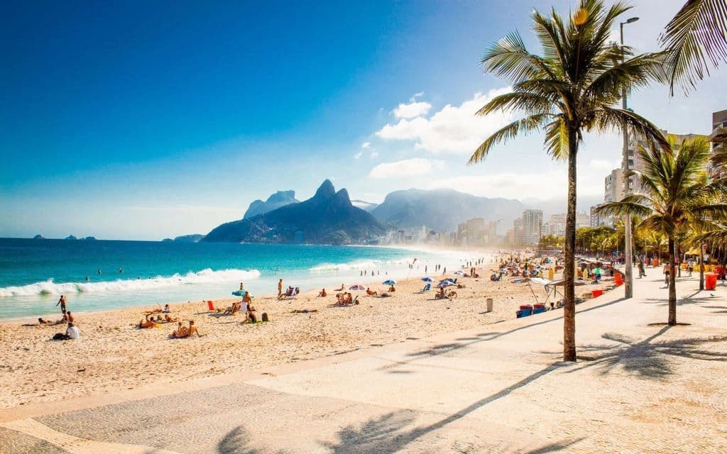 A wide view of a crowded tropical beach with people sunbathing, a city skyline, and mountains under a clear blue sky.