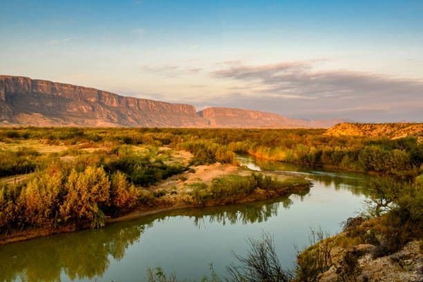 Un fiume sinuoso scorre attraverso una valle ricca di vegetazione ai piedi di un'ampia mesa al tramonto.