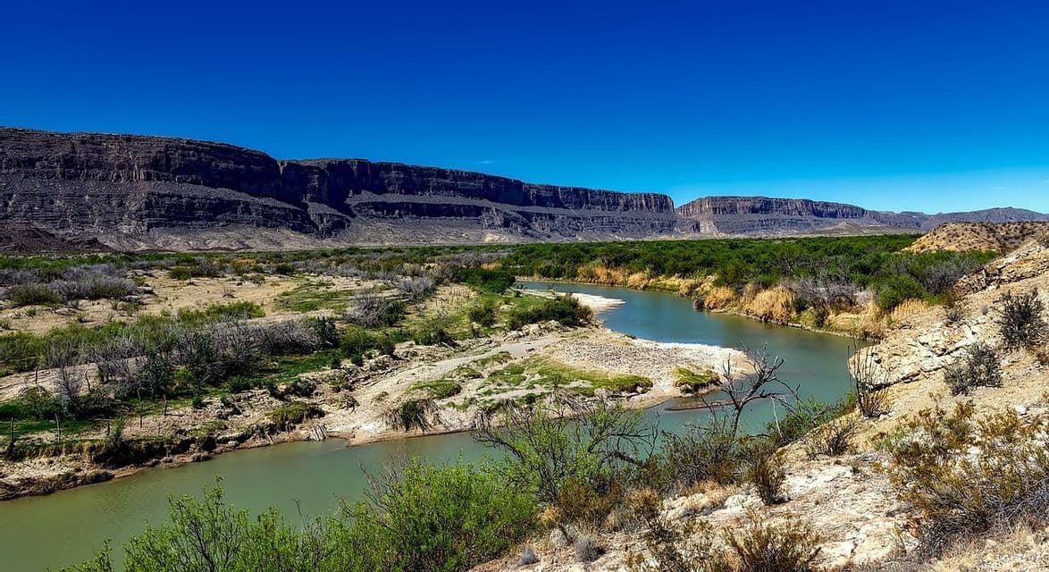 Un fiume serpeggia attraverso una valle desertica con canyon rocciosi sullo sfondo sotto un cielo azzurro chiaro.