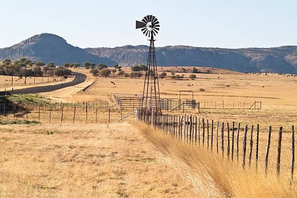 Un mulino a vento metallico si erge in un campo erboso e secco, accanto a una recinzione, con una strada asfaltata che si curva verso le montagne sullo sfondo.