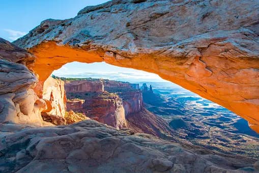 Un grande arco di pietra, illuminato dal sole, incornicia un'ampia vista di un paesaggio di canyon desertico sotto un cielo azzurro.