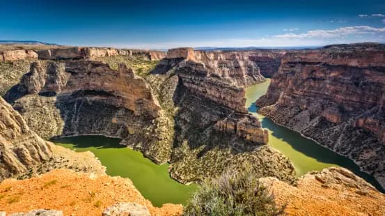 Un fiume verde si snoda in un vasto canyon roccioso sotto un cielo azzurro, visto da un punto panoramico elevato.