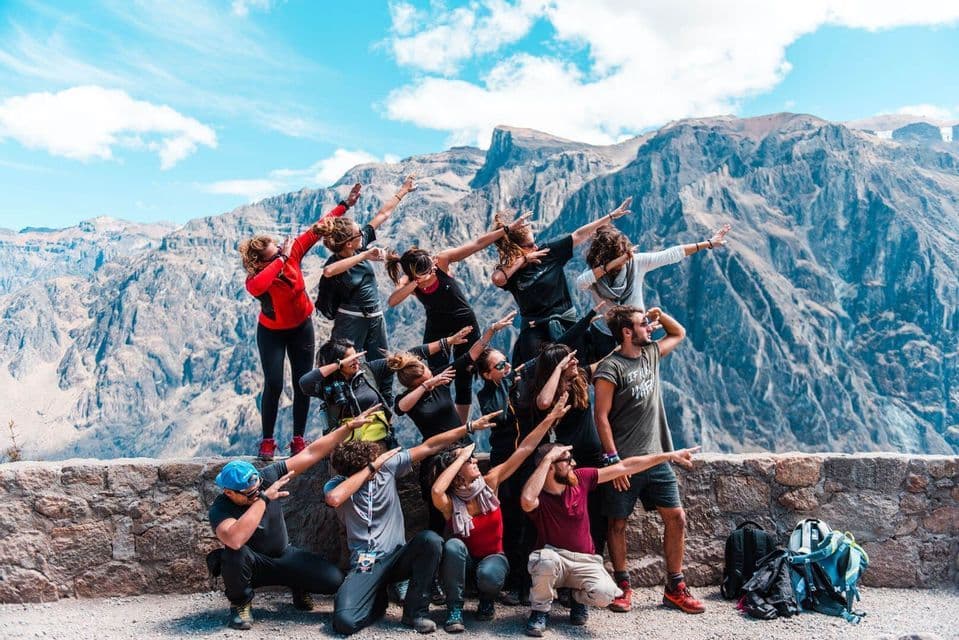 A WeRoad group trip does a synchronized 'dab' pose for a photo on a stone wall with a mountain range in the background.