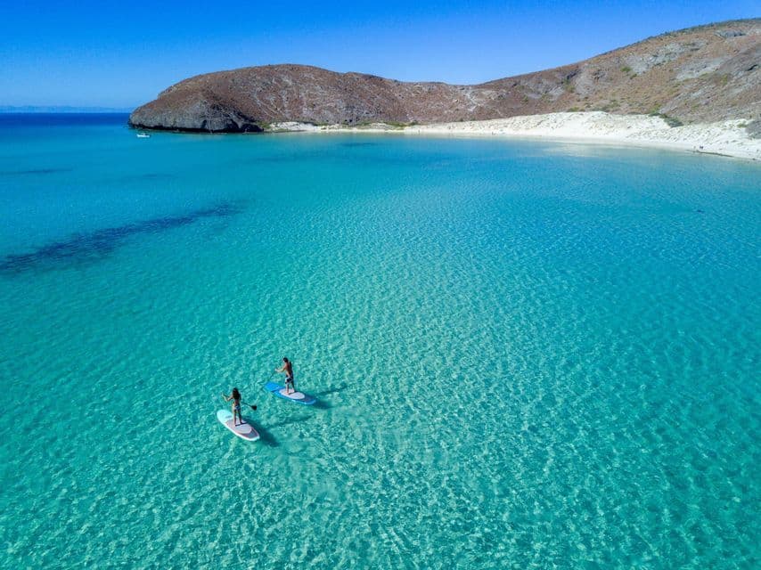 Vista aérea de dos personas en tablas de paddle surf en una bahía tranquila y cristalina de color turquesa, junto a una playa de arena y una colina árida.