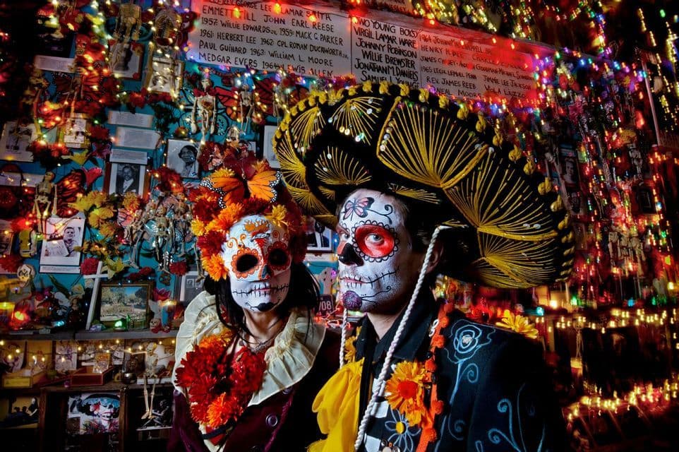 Un hombre y una mujer con maquillaje de calavera posan con trajes del Día de Muertos ante un altar colorido e iluminado.