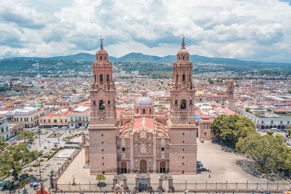 Una vista aérea de una gran catedral de piedra con dos torres, dominando una ciudad en expansión con montañas distantes bajo un cielo nublado.