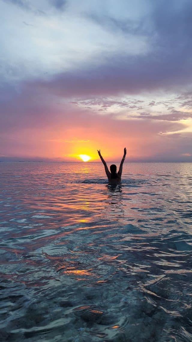 Une personne vue de dos se tient dans l'eau, les bras levés vers un coucher de soleil coloré à l'horizon.