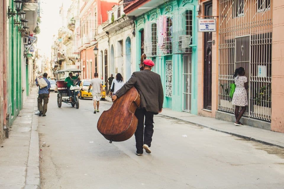 Un homme au bonnet rouge, vu de dos, porte une contrebasse en descendant une rue étroite bordée de bâtiments colorés.