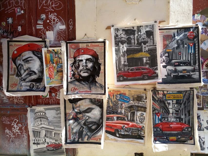 Una collezione di dipinti a tema cubano raffiguranti Che Guevara e auto d'epoca sono appesi a un muro coperto di graffiti.
