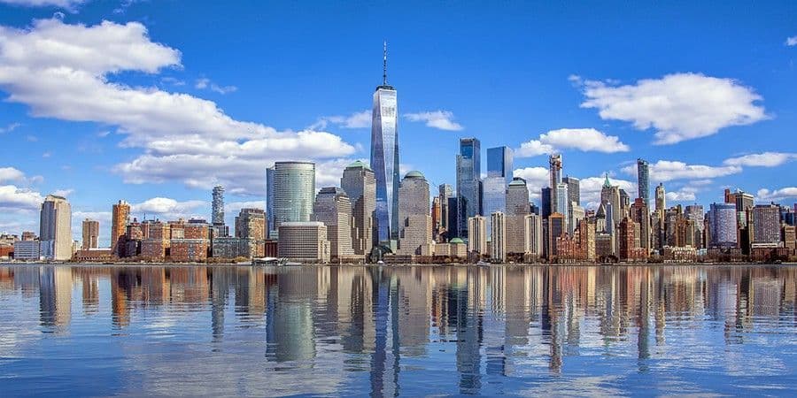 A modern city skyline with tall skyscrapers reflecting in the calm water under a blue sky with scattered clouds.