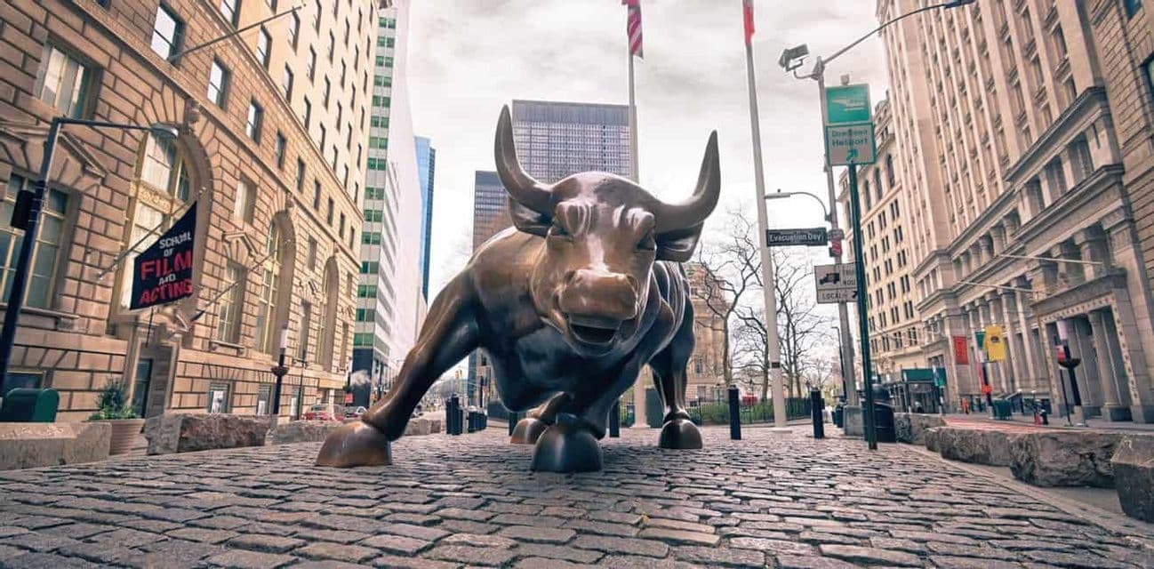 A low-angle view of the large bronze Charging Bull statue on a cobblestone street surrounded by tall city buildings.