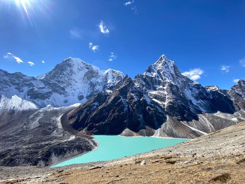 Un lago alpino de color turquesa brillante se asienta al pie de montañas escarpadas y cubiertas de nieve bajo un cielo azul soleado.