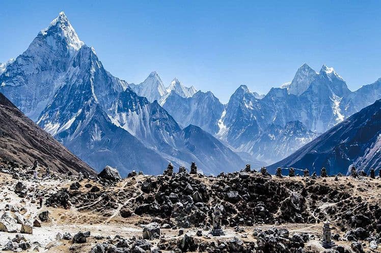 Una vasta cordillera cubierta de nieve se cierne sobre un paisaje rocoso y árido salpicado de pequeños monumentos de piedra bajo un cielo azul claro.