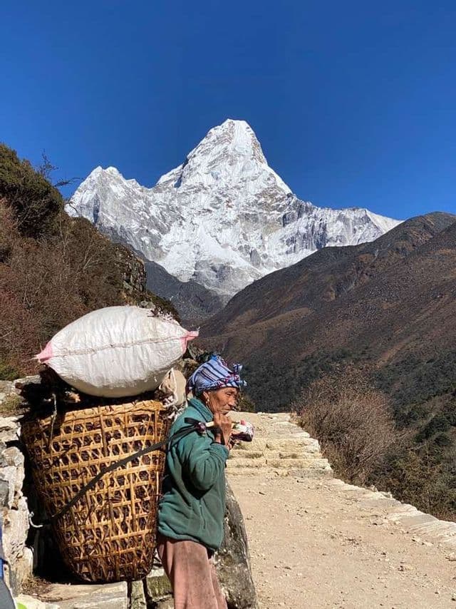Una persona con una gran cesta tejida a la espalda se detiene en un sendero de montaña con un pico nevado al fondo.