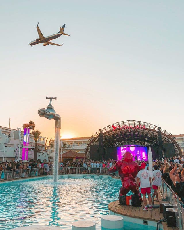 Ein tief fliegendes Flugzeug über einem überfüllten Pool-Musikfestival bei Sonnenuntergang, mit einer großen Bühne und einem riesigen Wasserhahn-Brunnen.