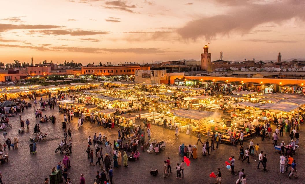 An elevated view of a bustling market square at sunset, filled with people walking between brightly lit stalls.
