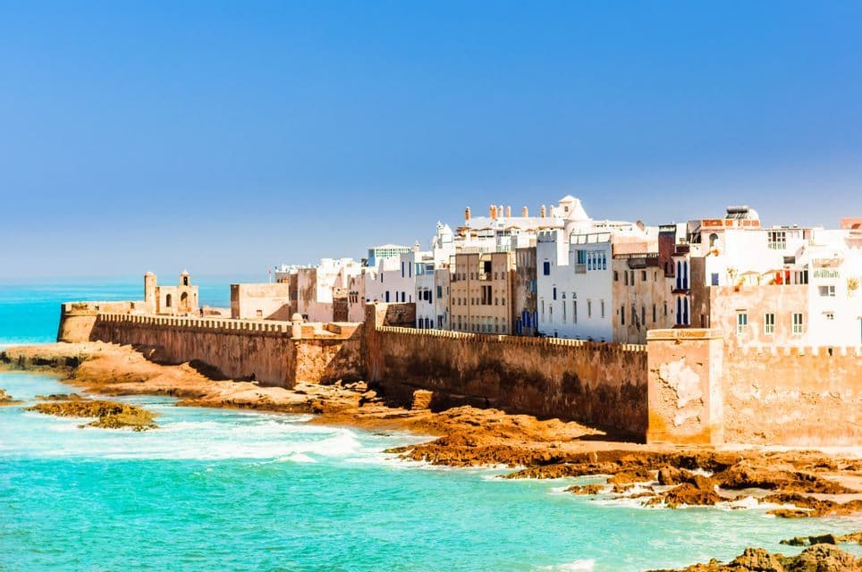 Una ciudad fortificada con edificios encalados en una costa rocosa con agua turquesa bajo un cielo despejado.