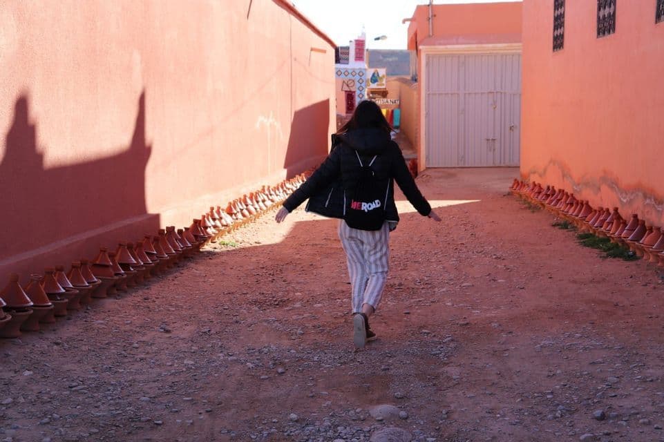 Eine Person mit WeRoad-Rucksack geht eine schmale Gasse entlang, mit Terrakotta-Mauern, gesäumt von einer Reihe Tajine-Töpfen.