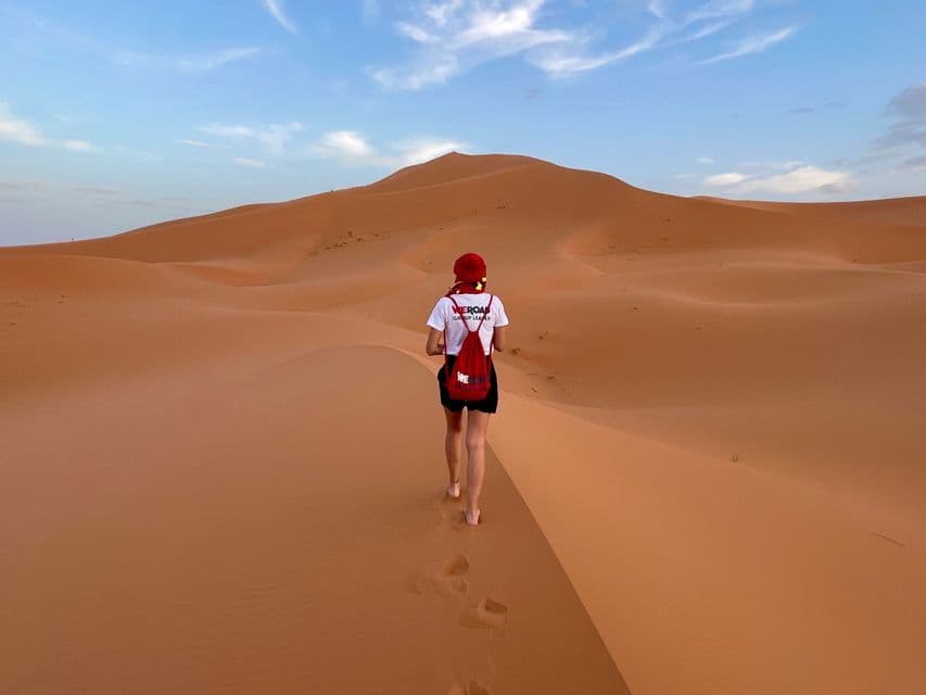 Un capogruppo WeRoad cammina a piedi nudi lungo la cresta di una vasta duna di sabbia arancione nel deserto, visto da dietro.