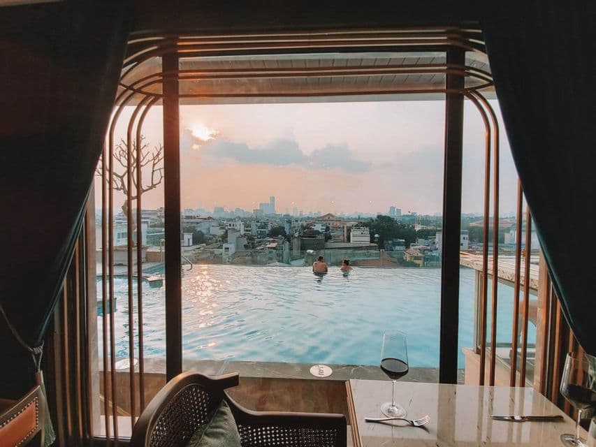 Dos personas en una piscina infinita en la azotea, con vistas a la ciudad al atardecer, observadas desde el interior de una habitación con una copa de vino sobre una mesa.