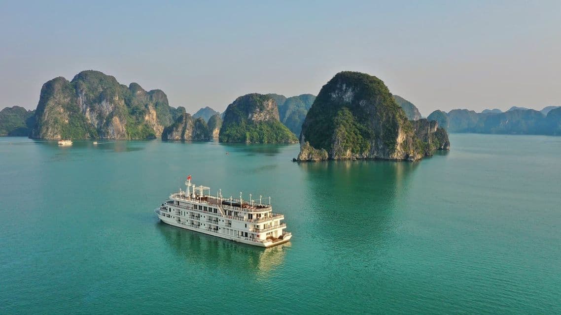 Un grand navire de croisière blanc navigue sur une eau turquoise calme parmi d'imposants karsts calcaires couverts de verdure.