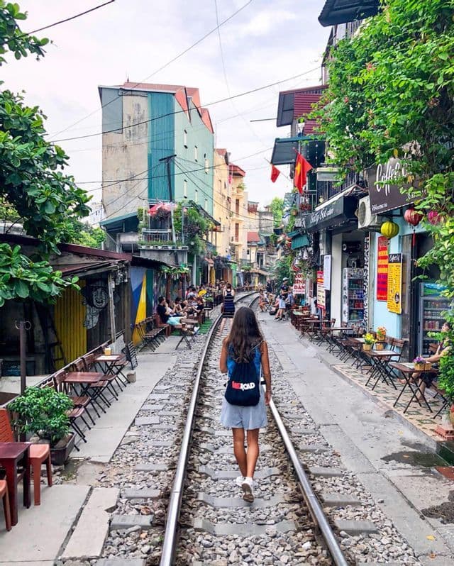 Une femme portant un sac à dos WeRoad se promène le long d'une voie ferrée qui traverse une rue étroite bordée de cafés en terrasse.